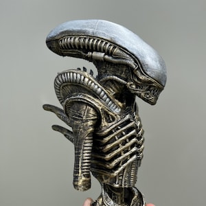 May include: A detailed bust of an Alien creature, featuring a silver head and a body with gold and bronze accents. The sculpture has intricate mechanical details and a textured finish, showcasing the iconic design from the film.