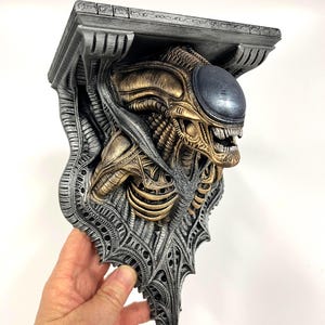 Puede incluir: Estantería detallada con un diseño de Xenomorfo. La estantería tiene una parte superior gris plateada y detalles intrincados, con la cabeza y la parte superior del cuerpo del alienígena en tonos dorados y plateados. La cabeza del alienígena tiene una cúpula gris azul oscuro.