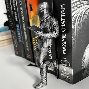 Peut inclure: Un serre-livres argenté en forme de personne en veste et pantalon, lisant un livre. Le serre-livres est appuyé contre une pile de livres avec des titres comme "SLEEP" et "STARFISH". Le serre-livres mesure environ 15 cm de haut.