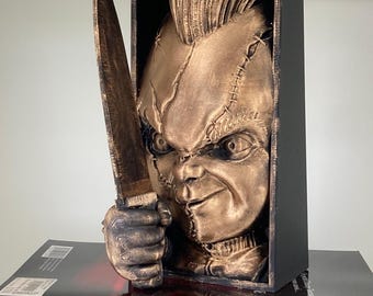May include: A bronze-colored 3D printed bust of a horror movie character with a stitched face and a mohawk hairstyle. The character is holding a knife and is framed in a black box.