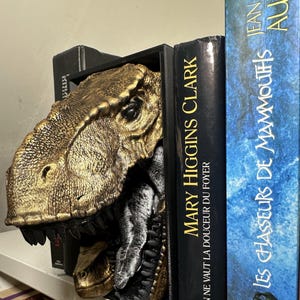 May include: A gold and black dinosaur head bookend. The T-Rex head is placed between two books, with the title "MARY HIGGINS CLARK" visible on one spine. The bookend provides a unique, decorative element to a bookshelf.