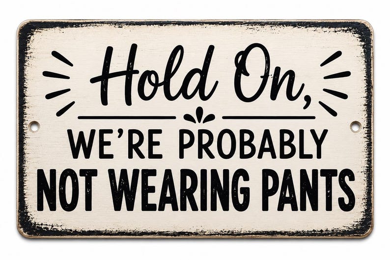 Op de afbeelding: Een rechthoekig gebroken wit metalen bord met versleten zwarte randen en de zwarte tekst Hold On, WE'RE PROBABLY NOT WEARING PANTS. Het bord heeft twee kleine ronde montagegaten aan de zijkanten en is voorzien van decoratieve zwarte lijnen.