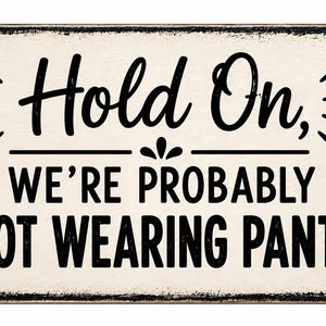 Op de afbeelding: Een rechthoekig bord met een verweerde zwarte rand en een gebroken witte achtergrond. De tekst luidt "Hold On, WE'RE PROBABLY NOT WEARING PANTS" in zwarte letters, met decoratieve lijnen en een klein bloemachtig accent tussen de regels.