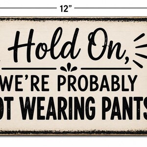 Op de afbeelding: Een rechthoekig metalen bord met een verweerde cr&egrave;mekleurige achtergrond en zwarte tekst die luidt: "Hold On, WE'RE PROBABLY NOT WEARING PANTS". Het bord heeft twee montagegaten aan de zijkanten en meet 30,48 cm breed bij 20,32 cm hoog.
