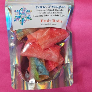 Op de afbeelding: Een zilveren hersluitbare zak met een wit label waarop staat "Little Freezies Freeze-Dried Candy. Fruits and Snacks Locally Made with Love Fruit Rolls 1.75 oz/49.62 grams". De zak bevat rode, roze en blauwe fruitrollen.
