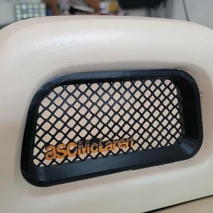 Può includere: Un componente automobilistico beige con una griglia nera e il testo arancione "asCmcclaren". Il componente ha una forma curva e arrotondata e sembra essere realizzato in plastica o materiale simile. Lo sfondo suggerisce un'officina.