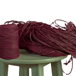 Può includere: Un rotolo di spago di colore bordeaux scuro si trova su uno sgabello verde. Lo spago è sciolto e sfilacciato, creando un mucchio disordinato.