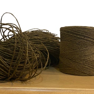 Può includere: Un rotolo di spago di carta marrone, un cordoncino in fibra naturale, è mostrato su una superficie di legno. Lo spago è avvolto strettamente ed è pronto per essere utilizzato per l'artigianato, l'imballaggio o il giardinaggio.
