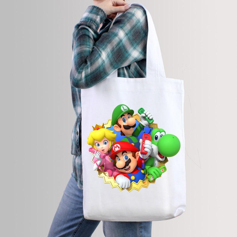 May include: White canvas tote bag with a colorful graphic print of Mario, Luigi, Peach, and Yoshi characters from the Super Mario video game series. The characters are holding controllers and are surrounded by a gold star.