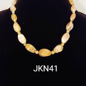May include: A necklace made of light brown and yellow oval-shaped stones. The necklace is on a black background. The text "JKN41" is printed below the necklace.
