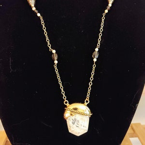 May include: A gold chain necklace with a white and gray stone pendant. The chain has small gold and black beads. The pendant is shaped like a teardrop and has a gold rim. The necklace is labeled "SBN1".