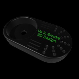 K&ouml;nnte beinhalten: Schwarzes, ovalf&ouml;rmiges 3D-gedrucktes Zubeh&ouml;r mit einer kreisf&ouml;rmigen Schale und einer Reihe kleiner L&ouml;cher und Linien. Der Text "Up In Smoke 3D Design" ist gr&uuml;n aufgedruckt.