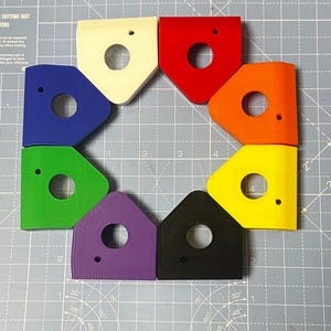 Könnte beinhalten: Eine Sammlung von acht farbenfrohen, geometrischen Formen, die kreisförmig angeordnet sind. Jedes Teil hat eine andere Farbe, darunter Weiß, Rot, Orange, Gelb, Grün, Blau, Lila und Schwarz. Jede Form hat ein kreisförmiges Loch und ein kleineres Loch.