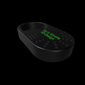 K&ouml;nnte beinhalten: Schwarzer Aschenbecher mit einer kreisf&ouml;rmigen Halterung und einer rechteckigen Basis. Der Aschenbecher hat eine Reihe kleiner L&ouml;cher und den Text "Up In Smoke 3D Design" in Gr&uuml;n. Der Aschenbecher ist f&uuml;r Rauchwaren konzipiert.