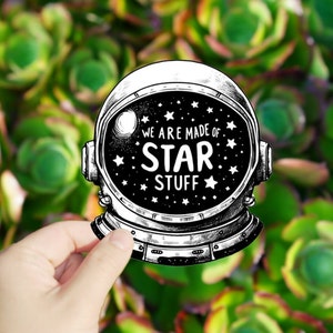 May include: A black and white illustration of an astronaut's helmet with the text "We are made of star stuff" inside. The helmet is held in a hand against a green background.