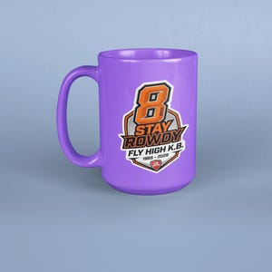 May include: A purple ceramic mug featuring a graphic design with a large orange number 8, the text STAY ROWDY, FLY HIGH K.B., and the years 1985-2026. The design is enclosed in a shield-shaped emblem with black, white, and orange accents.