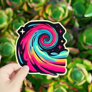 May include: A colorful sticker with a swirling galaxy design. The galaxy is pink, blue, and yellow with a black background. There are small white stars scattered throughout the galaxy.
