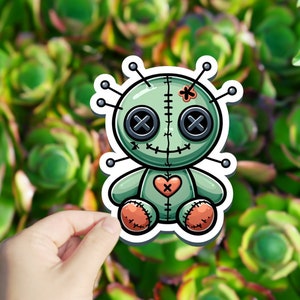 May include: A green voodoo doll with a stitched-on heart, black button eyes, and a red X on its chest. The doll has a white outline and is held in a hand against a green background.