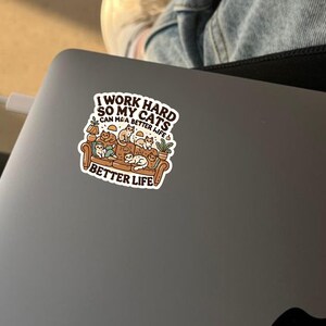 May include: A sticker with a cartoon illustration of a couch full of cats. The text reads "I work hard so my cats can have a better life."  The sticker is on a gray laptop.