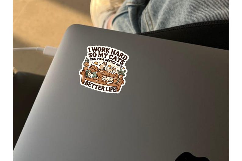 May include: A sticker with a cartoon illustration of a couch full of cats. The text reads "I work hard so my cats can have a better life."  The sticker is on a gray laptop.