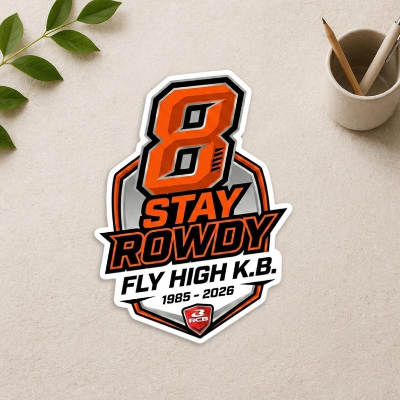 May include: A die-cut sticker featuring a large orange number 8 above the text STAY ROWDY and FLY HIGH K.B. with the years 1985-2026. The design includes a shield-shaped background in grey and orange with a small red RCB logo at the bottom center.