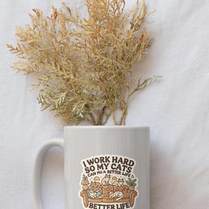 May include: White ceramic mug with a brown and white sticker that says "I work hard so my cats can have a better life" with a cartoon image of cats on a couch.