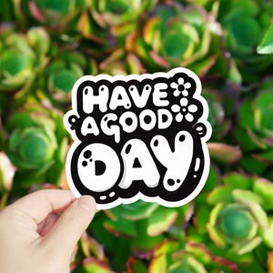 May include: A white sticker with black lettering that says "Have a good day" with a flower design. The sticker is being held in a hand against a background of green plants.