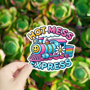 May include: A colorful cartoon train sticker with the text "Hot Mess Express". The train is blue, pink, and yellow with a smiley face on the front. The sticker is on a green background.