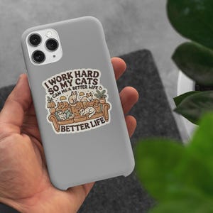 May include: A white phone case with a colorful illustration of cats sitting on a couch. The text on the case reads "I work hard so my cats can have a better life".