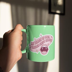 May include: A light green ceramic mug with a handle. The mug features a sticker with the text "MY BRAIN IS 90% TAYLOR SWIFT LYRICS" in a pink speech bubble with a cartoon mouth graphic. The mug is held by a person.
