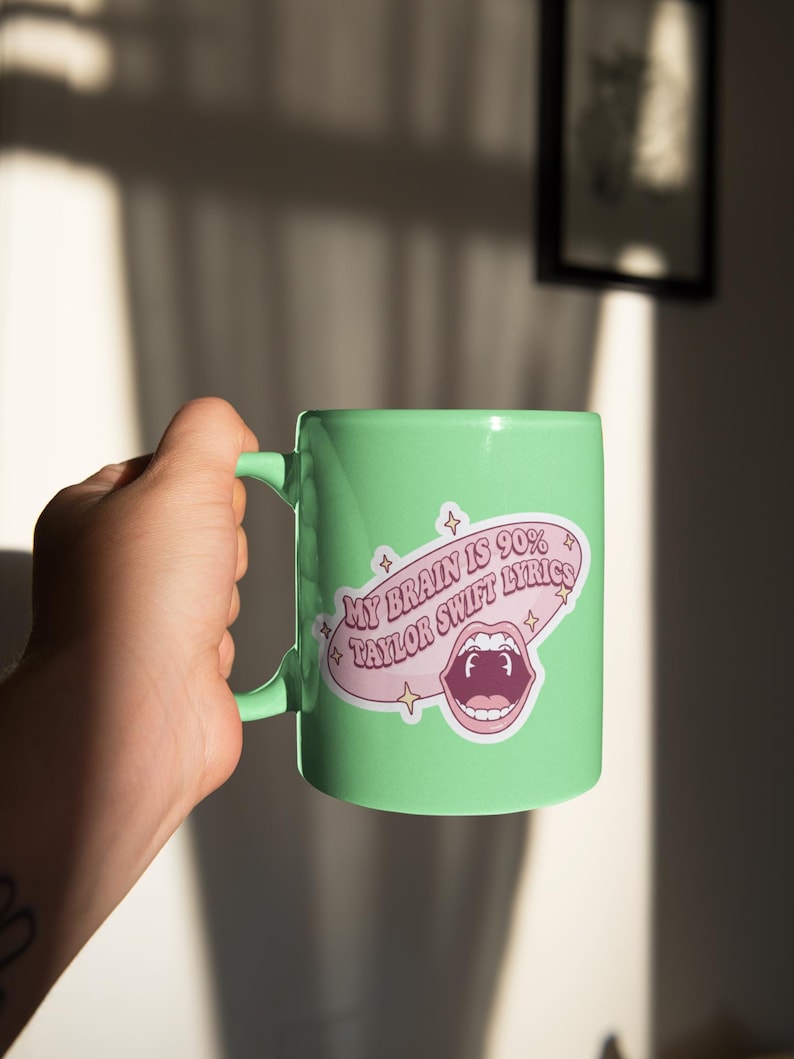 May include: A light green ceramic mug with a handle. The mug features a sticker with the text "MY BRAIN IS 90% TAYLOR SWIFT LYRICS" in a pink speech bubble with a cartoon mouth graphic. The mug is held by a person.