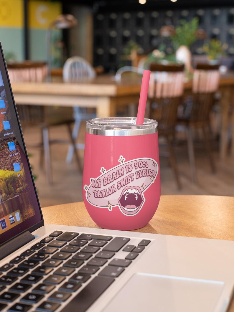 May include: A pink insulated wine tumbler with a silver rim and a pink straw. The tumbler has a sticker that reads "My Brain is 90% Taylor Swift Lyrics" with an open mouth graphic.