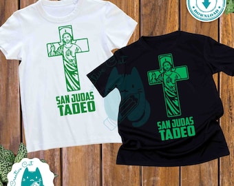 May include: Two t-shirts, one white and one black, each featuring a green graphic of a cross with an image of a religious figure and the text "SAN JUDAS TADEO". The shirts are displayed on a wooden surface.