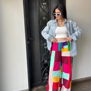 May include: A woman wearing a white tank top, a light blue denim jacket, and colorful patchwork wide-leg pants. She is wearing white sunglasses and white sneakers.