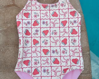 Peut inclure: Un maillot de bain une pièce avec un motif de fraises. Le tissu blanc est orné de fraises roses, de nœuds et de fleurs dans une grille rouge. Les bretelles sont nouées aux épaules avec des nœuds roses, et l'intérieur est rose uni.