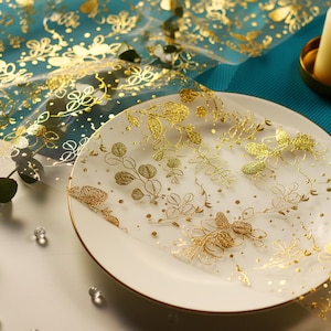 Peut inclure: Une assiette blanche avec un bord doré est partiellement recouverte d'un voile transparent à motifs de feuilles d'or. Le voile présente des motifs floraux et pointillés délicats. L'arrière-plan comprend une surface turquoise et du tissu à feuilles d'or.