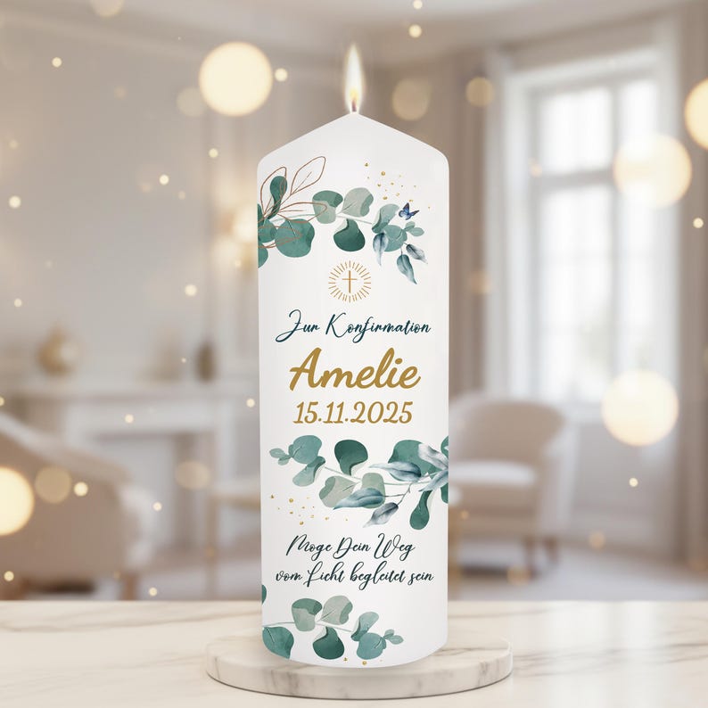 May include: A white pillar candle with a lit flame. The candle is decorated with green and blue eucalyptus leaves and the name "Amelie" in gold script. The text "Zur Konfirmation" and a date are also visible.