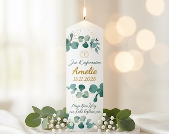 May include: A white pillar candle with a lit flame, adorned with green eucalyptus leaves and gold text. The candle is personalized with a name and date, and rests on a white marble tray with greenery.