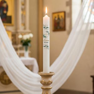 May include: A lit white candle with a floral design and the name "Laura" printed on it. The candle is in a decorative holder. The background includes a church interior with white drapes.