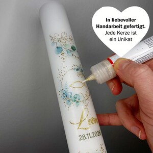 May include: White candle decorated with blue and gold floral designs, the name "Leon" and the date 28.11.2026 in gold script. The candle is being embellished with gold glitter. Text on the image reads "In liebevoller Handarbeit gefertigt. Jede Kerze ist ein Unikat."