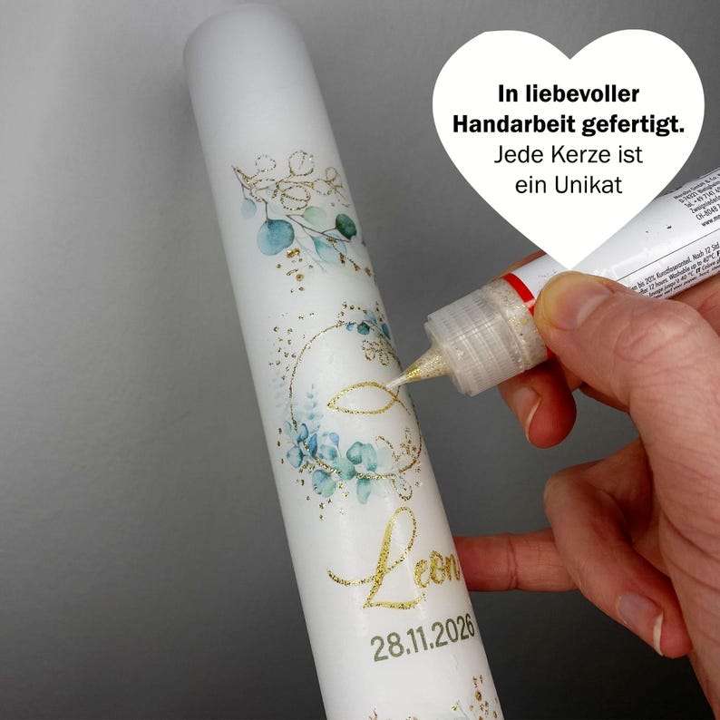 May include: White candle decorated with blue and gold floral designs, the name "Leon" and the date 28.11.2026 in gold script. The candle is being embellished with gold glitter. Text on the image reads "In liebevoller Handarbeit gefertigt. Jede Kerze ist ein Unikat."