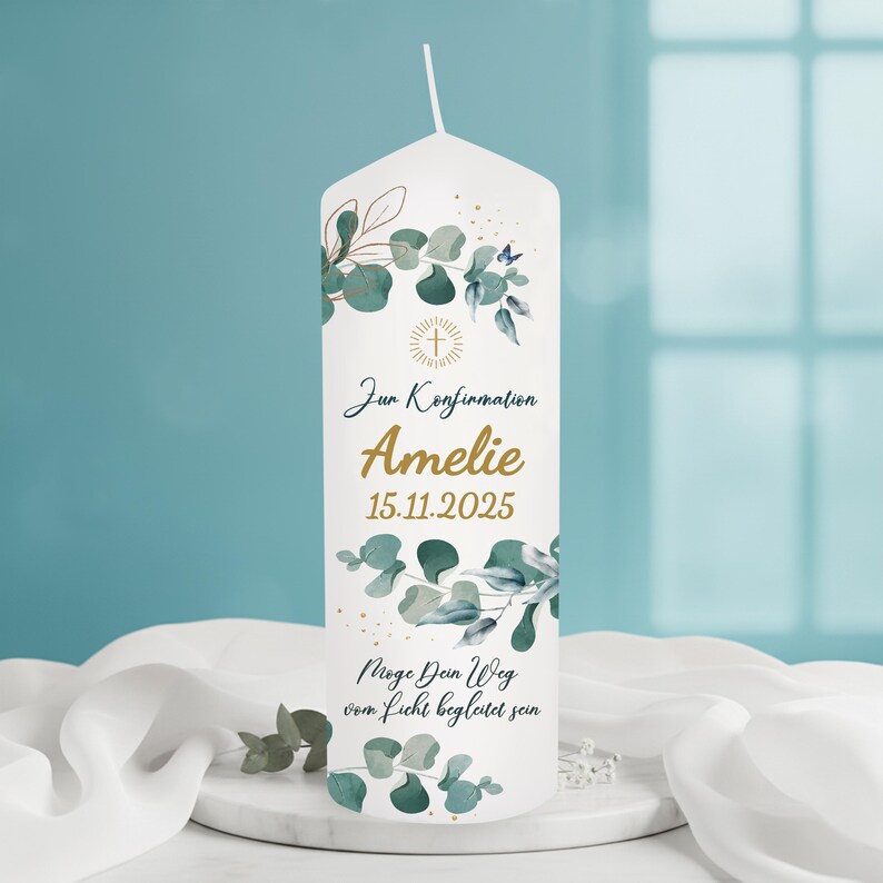 May include: A white pillar candle decorated with green and blue eucalyptus leaves and the text "Zur Konfirmation Amelie 15.11.2025". The candle also features a cross and the phrase "M&ouml;ge Dein Weg vom Licht begleitet sein".