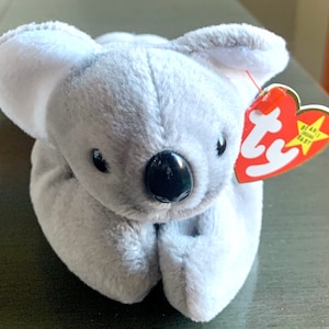 May include: A gray plush koala bear with black eyes and a black nose. The bear is lying on its belly with its paws tucked under its chin. The bear has a red Ty tag attached to its ear with the words "Beanie Babies" printed on it.