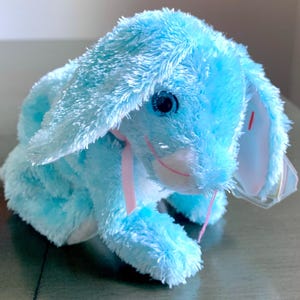 May include: A light blue plush bunny with a pink ribbon around its neck. The bunny has a white face with a pink nose and blue eyes.