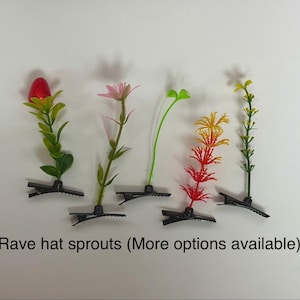 以下が含まれることがあります： 赤い蕾、ピンクの花、緑の芽、黄色とオレンジの羽のような植物、細い緑の茎など、様々な植物や花のモチーフが付いた5つのヘアクリップ。下のテキストには「Rave hat sprouts (More options available)」と書かれています。