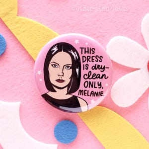 May include: A circular pin-back button with a pink background featuring a stylized illustration of a person with dark hair and the black text: THIS DRESS IS dry-clean ONLY, MELANIE. Small white stars are scattered around the illustration.