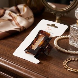 Peut inclure: Une pince à cheveux en écaille de tortue de forme rectangulaire, présentée sur une carte blanche avec le mot "Diva". La pince est marron et présente un design géométrique. Un collier de perles et un bracelet doré sont également visibles.