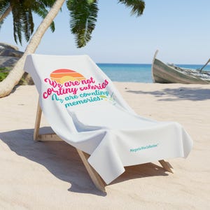 May include: A white beach towel draped over a wooden lounge chair. The towel features a graphic of a sunset and a cocktail glass with the text: We are not counting calories, we are counting memories. The text Margarita Rita Collection is printed.