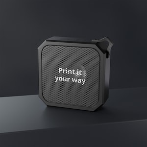 May include: A black, square-shaped device with a perforated front panel. The text "Print it your way" is displayed in white, illuminated letters on the front. The device has a sleek, modern design.