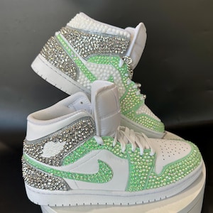 May include: White high-top sneakers with green and silver rhinestones. The sneakers have a white swoosh logo and a white sole.
